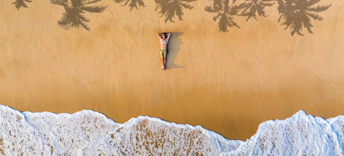 Drohnenaufnahme eines Mannes, der entspannt auf dem Rücken im Sand eines goldgelben Strandes liegt. Die Brandung rollt sanft an den unteren Bildrand, während sich die Schatten von Palmen auf den Sand zeichnen. Die Szene wirkt ruhig, tropisch und friedlich