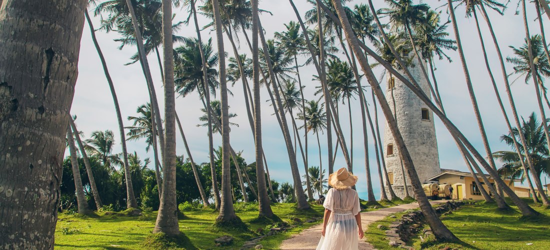 Frau in weißem Kleid und Strohhut spaziert durch Palmen zum historischen Leuchtturm von Galle in Sri Lanka