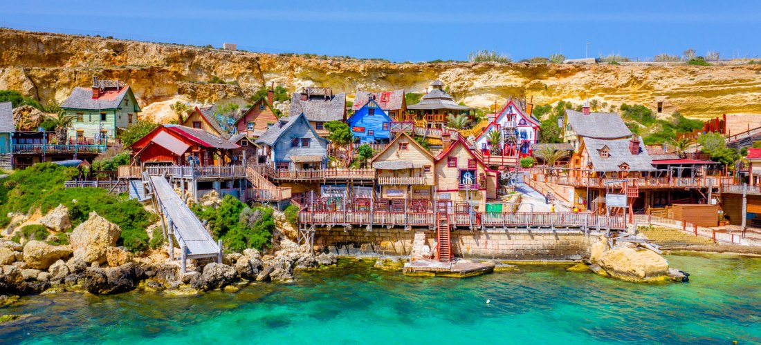 Bunte Holzhäuser des Popeye Village auf Malta an der Anchor Bay mit türkisblauem Wasser und Felsküste im Hintergrund