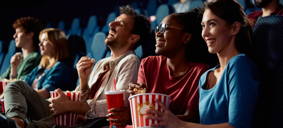 Mehrere junge Menschen sitzen im Kino, lachen und genießen Popcorn und Getränke. Die Stimmung ist ausgelassen und fröhlich, die Sitze sind blau und der Raum leicht abgedunkelt.