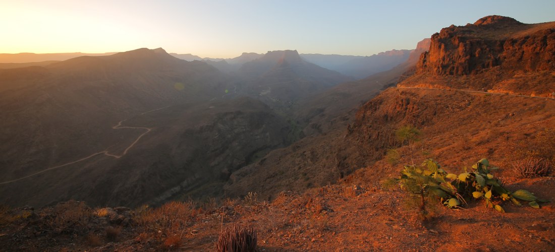 Sonnenuntergang über dem Fataga-Tal auf Gran Canaria mit roter Felslandschaft, kurviger Straße und Bergen im Hintergrund