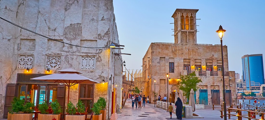 Dubai - Creek Szene in der historischen Altstadt von Dubai bei Sonnenuntergang. Traditionelle Lehmgebäude mit kunstvoll verzierten Fenstern und einem typischen Windturm säumen die Fußgängerzone. Menschen flanieren zwischen kleinen Cafés, Lichterketten und Straßenlatern