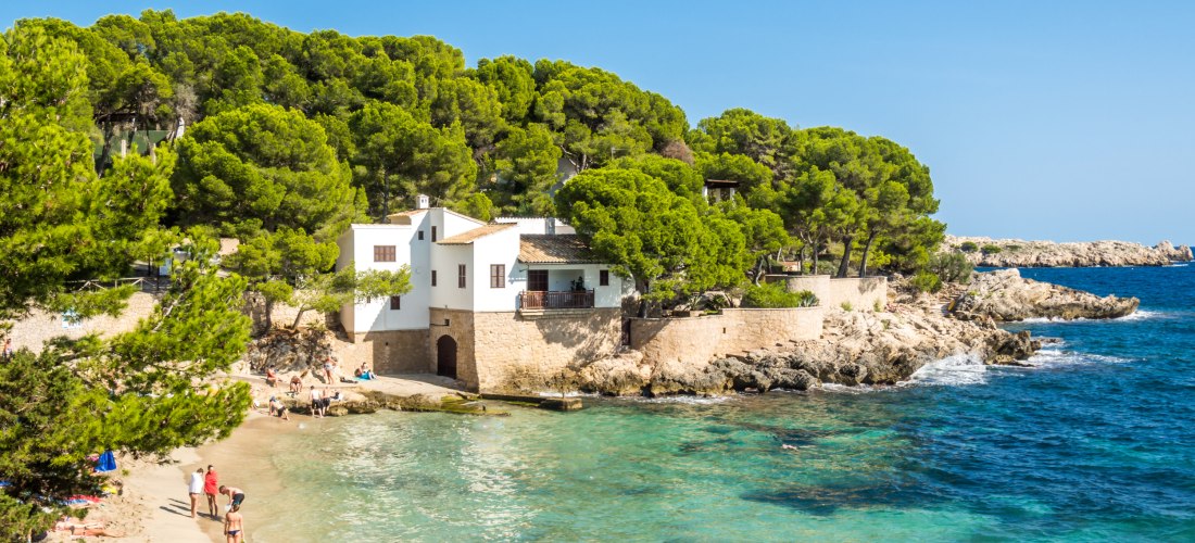 Kleine Badebucht mit weißem Haus und grünen Pinien bei Cala Ratjada auf Mallorca, Menschen genießen das klare Meer und den Strand