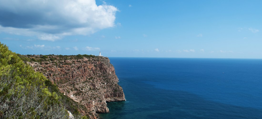 Steilküste mit rotbraunen Felsen und grüner Vegetation auf Formentera, im Hintergrund ein weißer Leuchtturm auf einer Klippe. Unten breitet sich das tiefblaue Mittelmeer aus, am Himmel ziehen weiße Wolken vorbei.