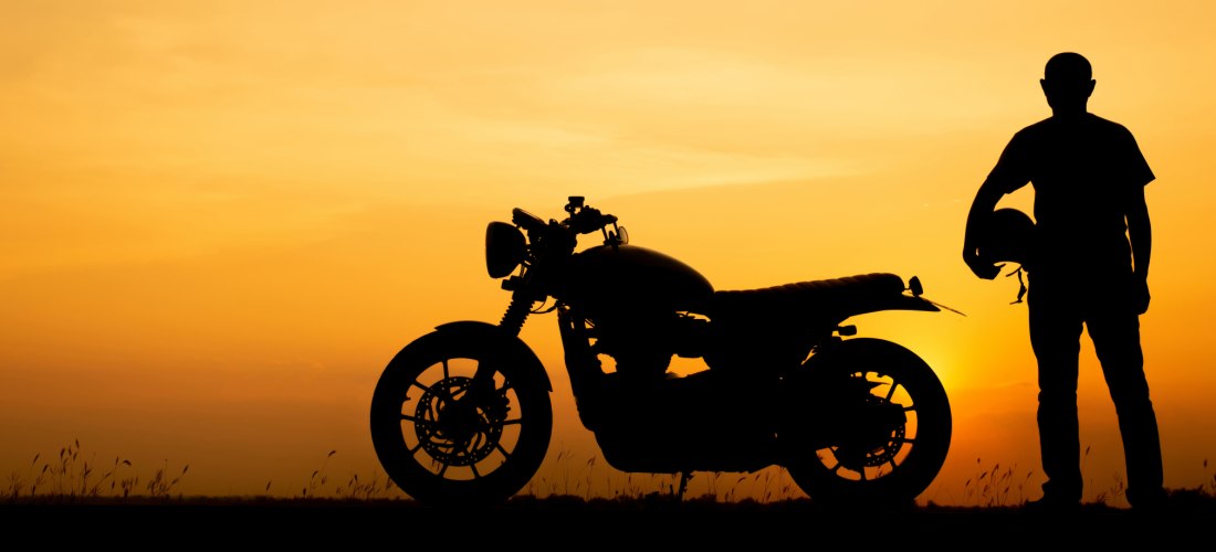 Schattenriss eines Motorradfahrers mit Helm in der Hand neben einem klassischen Motorrad bei Sonnenuntergang. Der Himmel leuchtet in warmen Orange- und Gelbtönen, während die Szene Ruhe und Abenteuerlust zugleich ausstrahlt.