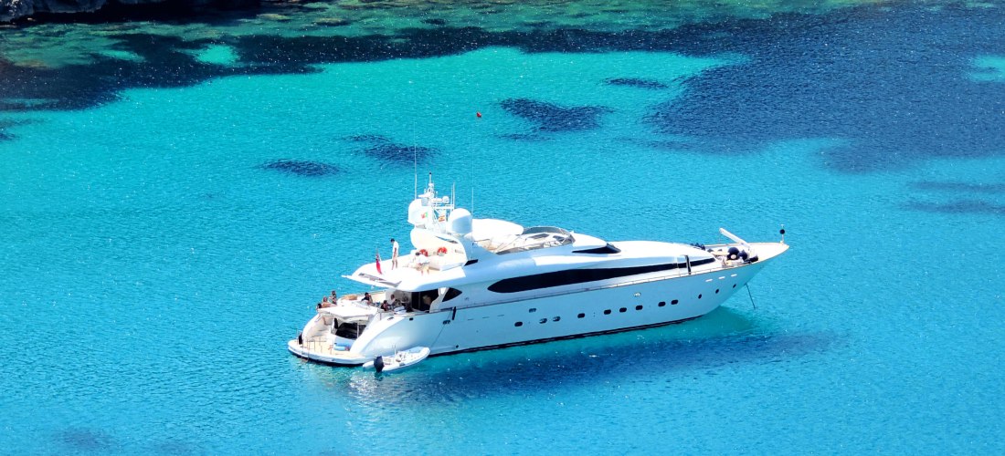 Weiße Luxus-Yacht ankert in einer traumhaft klaren, türkisblauen Bucht mit felsigem Ufer. Das Wasser ist so klar, dass dunklere Unterwasserformationen sichtbar sind. Mehrere Personen entspannen auf Deck und genießen die Sonne. Die Szenerie wirkt exklusiv 