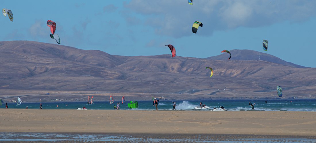Weitläufiger Sandstrand mit vielen Kitesurfern und bunten Kites in der Luft. Im Hintergrund ragen braune Hügel unter blauem Himmel auf