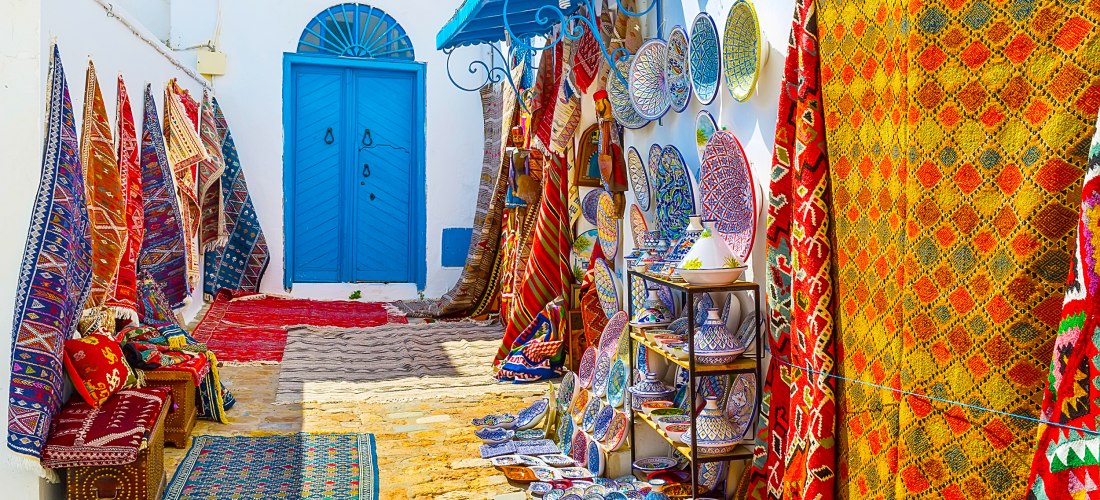 Eine enge Gasse in Sidi Bou Said, Tunesien, gesäumt von weißen Hauswänden mit einer strahlend blauen Tür. Links und rechts der Gasse hängen bunte Teppiche und Decken, die die Straße in ein Farbenmeer verwandeln. Dazwischen sind Töpferwaren und traditionel