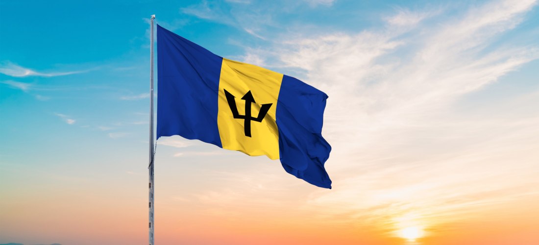Die Nationalflagge von Barbados weht im Wind vor einem farbenprächtigen Sonnenuntergang über dem Meer. Die Flagge besteht aus drei vertikalen Streifen – zwei blaue Streifen außen und ein goldener Streifen in der Mitte mit einem schwarzen Dreizack-Symbol d
