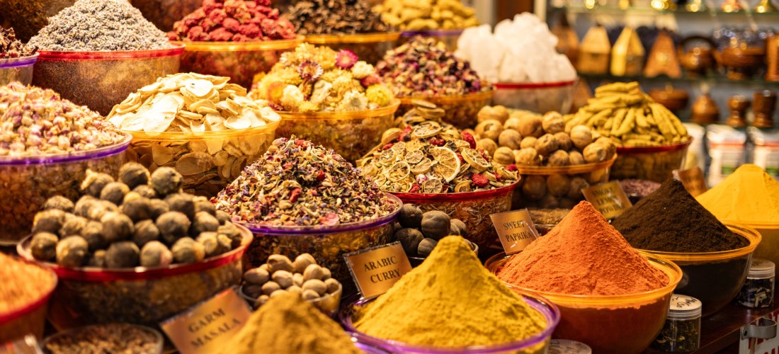 Dubai - Deira Bunte Auswahl an Gewürzen und getrockneten Kräutern in einem arabischen Souk. Die Gewürze sind in offenen Schalen gestapelt, darunter Kurkuma, Paprika, Zimt, arabisches Curry und getrocknete Zitronen. Im Hintergrund stehen kunstvoll dekorierte Teegläser u