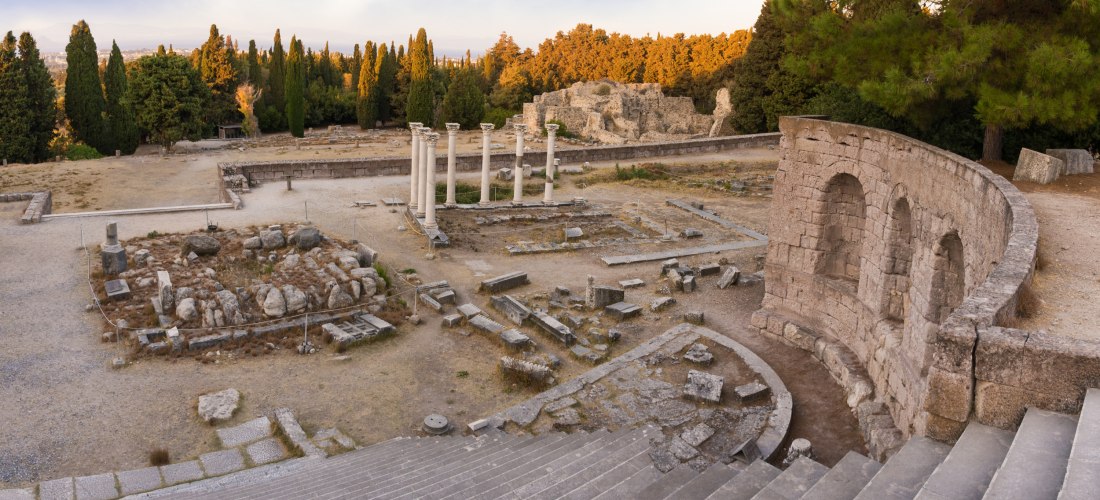 Antikes halbrundes Theater mit Sitzstufen, daneben Tempelruinen mit mehreren stehenden Säulen, umgeben von Bäumen und Grabungsfeld