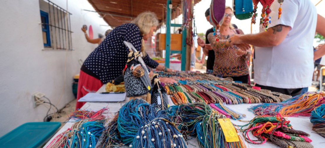 Marktstand mit einer großen Auswahl an handgefertigten, farbenfrohen Armbändern und Schmuck aus Garn. Menschen betrachten die Waren, ein Strohdach spendet Schatten