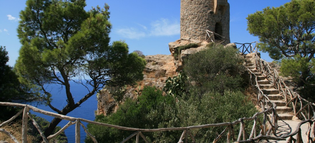 Das Bild zeigt eine alte Wachturm-Ruine auf einer Felskuppe hoch über dem Meer. Der Turm ist rund, aus groben Natursteinen gebaut, mit kleinen Fenstern und einer hölzernen Balustrade, die sich den Felsen entlang schlängelt. Der Himmel ist strahlend blau, 