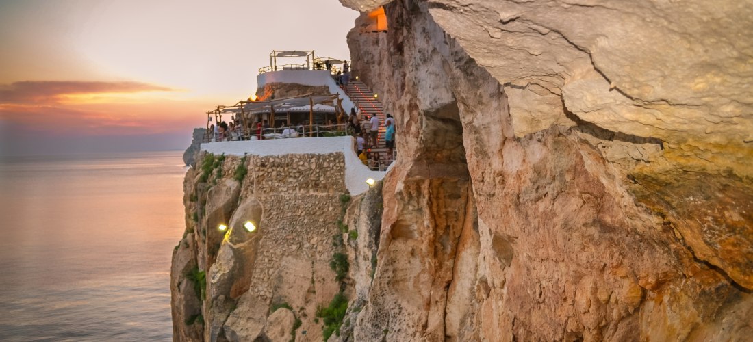 Eine Felsenbar auf Menorca, spektakulär in die steile Klippe eingelassen, bei Sonnenuntergang. Die Bar liegt auf einer Plattform mit Geländern und gemütlicher Einrichtung, Besucher genießen den Ausblick auf das ruhige Meer. Der Himmel ist orange-violett g