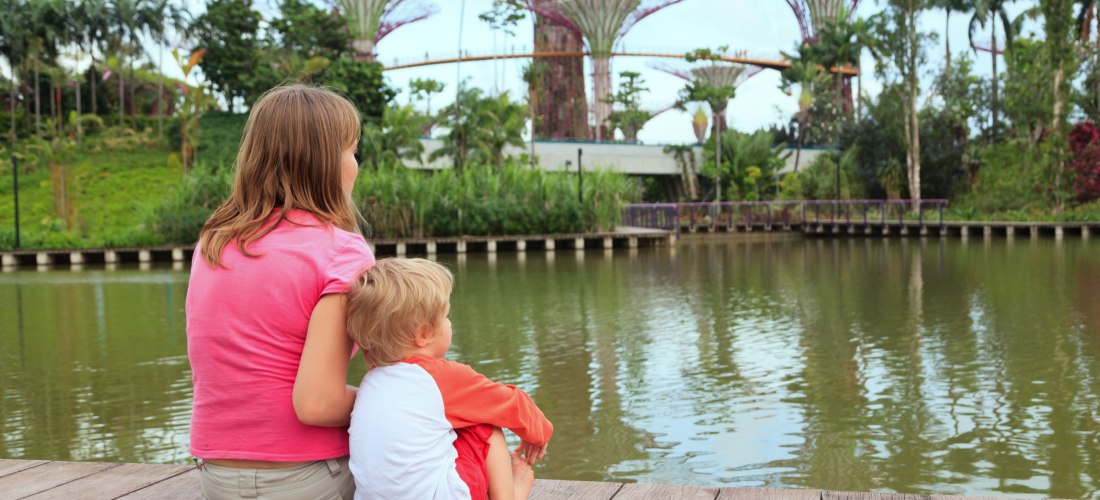 Ein Mädchen und ein kleiner Junge sitzen auf einem Holzsteg an einem ruhigen See in Singapur. Im Hintergrund sind die futuristischen Supertrees von Gardens by the Bay zu sehen. Die Kinder blicken ruhig auf das Wasser und die üppige tropische Vegetation.