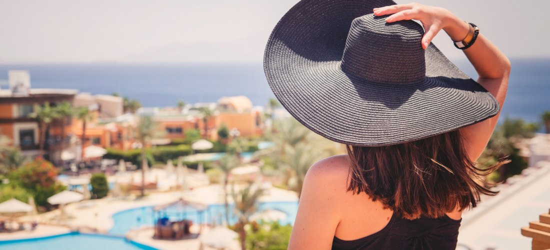 Eine Frau mit dunklem Haar und großem Sonnenhut blickt von einem Balkon auf ein weitläufiges Resort mit Pools, Palmen und orangefarbenen Gebäuden. Im Hintergrund ist das tiefblaue Meer zu sehen. Die Szene wirkt entspannt und sommerlich.