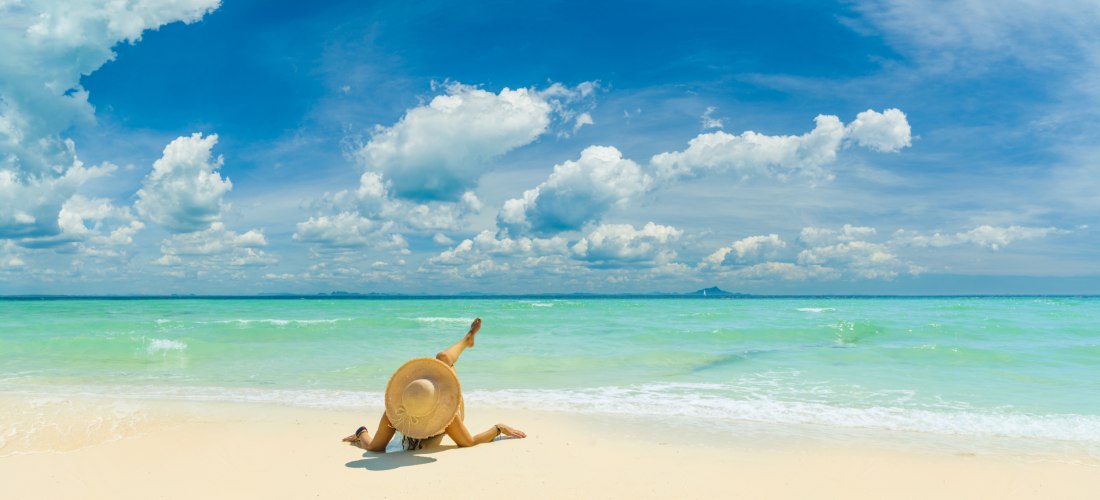 eine Frau mit Sonnenhut liegt entspannt am weißen Sandstrand mit türkisfarbenem Meer und blauem Himmel