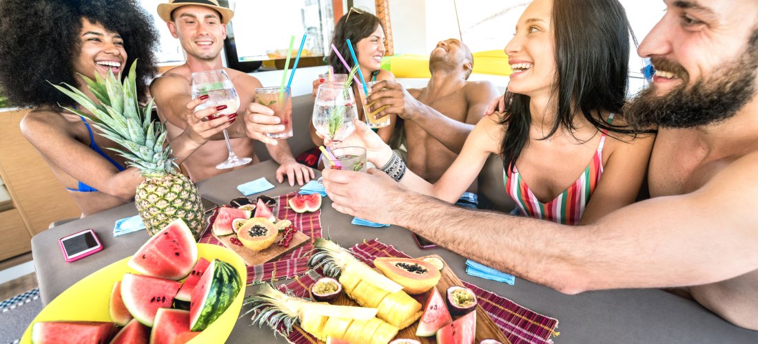 Gruppe junger Menschen in Badekleidung sitzt lachend an einem Tisch auf einem Boot oder in einer Strandlounge. Sie stoßen mit Cocktails an, auf dem Tisch liegen tropische Früchte wie Wassermelone, Ananas, Papaya und Maracuja. Die Stimmung ist ausgelassen 