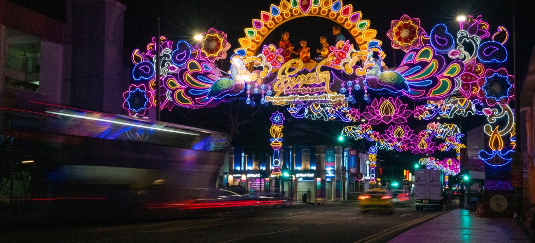 Leuchtend bunte Deepavali-Straßendekorationen in Singapur bei Nacht. Die Straße ist mit aufwendigen, farbenfrohen Lichtinstallationen überspannt, die Pfauen, Blumen und hinduistische Figuren zeigen. Im Vordergrund fahren Autos und ein Doppeldeckerbus durc