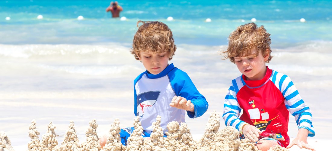 Zwei kleine Jungen mit lockigen Haaren sitzen am weißen Sandstrand und bauen konzentriert eine fantasievolle Sandburg mit tropfenartigen Türmchen. Der Junge links trägt ein blau-weißes UV-Shirt mit Haifisch-Motiv, der rechte ein rot-blaues Shirt mit Pirat