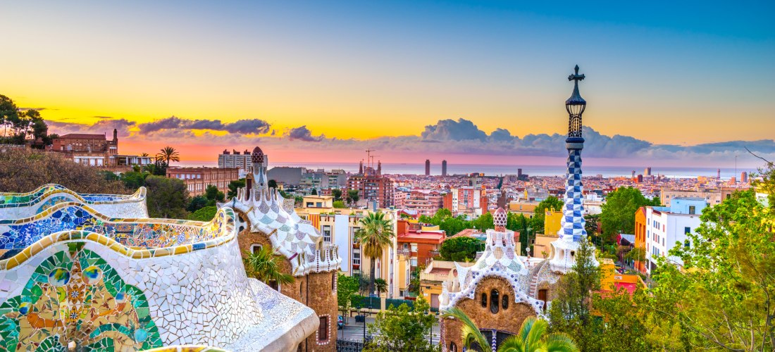 Blick über Barcelona vom Park Güell aus bei Sonnenuntergang. Im Vordergrund bunte, mosaikverzierte Architektur von Antoni Gaudí, im Hintergrund die Skyline der Stadt und das Mittelmeer unter einem farbenfrohen Himmel.