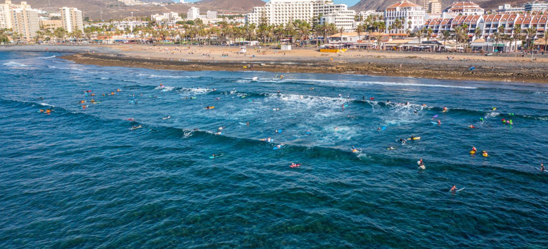 Surfer im Meer, dahinter breiter Sandstrand, Promenade mit Palmen und Hotels vor den Bergen.