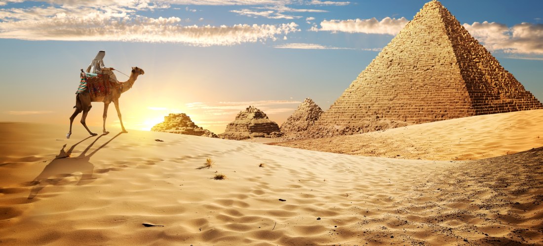 Das Bild zeigt eine weite Wüstenlandschaft bei Sonnenuntergang. Im Vordergrund zieht ein Mann in traditioneller arabischer Kleidung auf einem Kamel durch den weichen, goldfarbenen Sand. Die Sonne geht hinter ihm unter und wirft lange Schatten. Im Hintergr