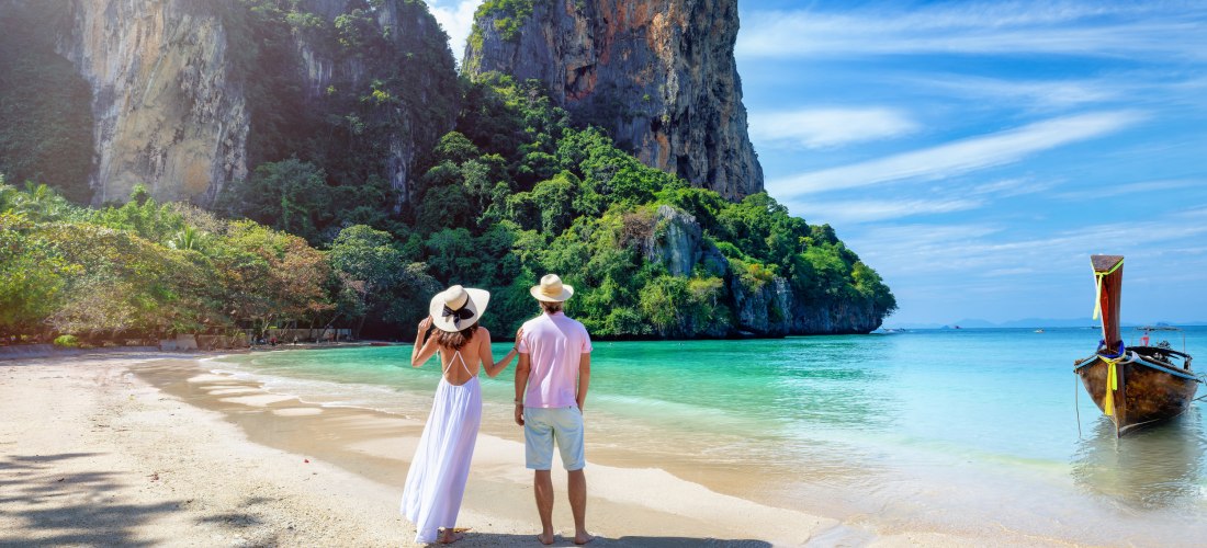 Das Bild zeigt ein Paar in sommerlicher Kleidung, das entspannt an einem tropischen Sandstrand steht. Die Frau trägt ein weißes, luftiges Kleid und einen Sonnenhut, der Mann helle Shorts, ein rosa Shirt und ebenfalls einen Hut. Beide blicken auf das ruhig