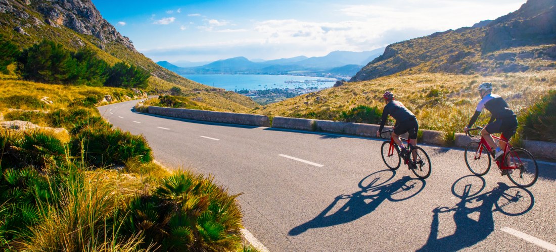 Zwei Rennradfahrer fahren auf einer kurvigen, asphaltierten Panoramastraße durch eine bergige Landschaft mit Blick auf eine Bucht. Die Umgebung ist geprägt von goldenen Gräsern, Felsen und einzelnen Büschen. Im Hintergrund erstreckt sich das Meer mit eine