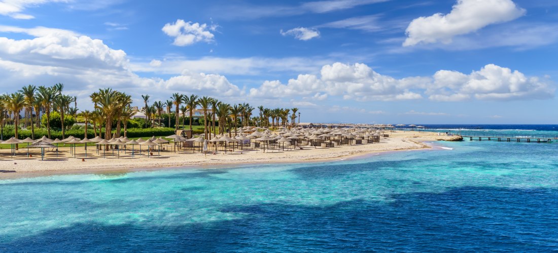 Langer Sandstrand mit vielen Sonnenschirmen und Palmen. Das Meer leuchtet türkis und blau, ein Steg führt ins Wasser. Am Horizont ziehen sich weiße Wolken über den klaren Himmel