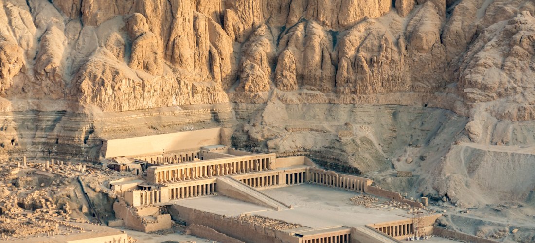 Das Bild zeigt eine weite Wüstenlandschaft mit einem monumentalen, mehrstufigen Terrassentempel, dem Totentempel der Pharaonin Hatschepsut in Deir el-Bahari. Der Tempel besteht aus langen Säulengängen, breiten Rampen und offenen Höfen. Er ist direkt in ei