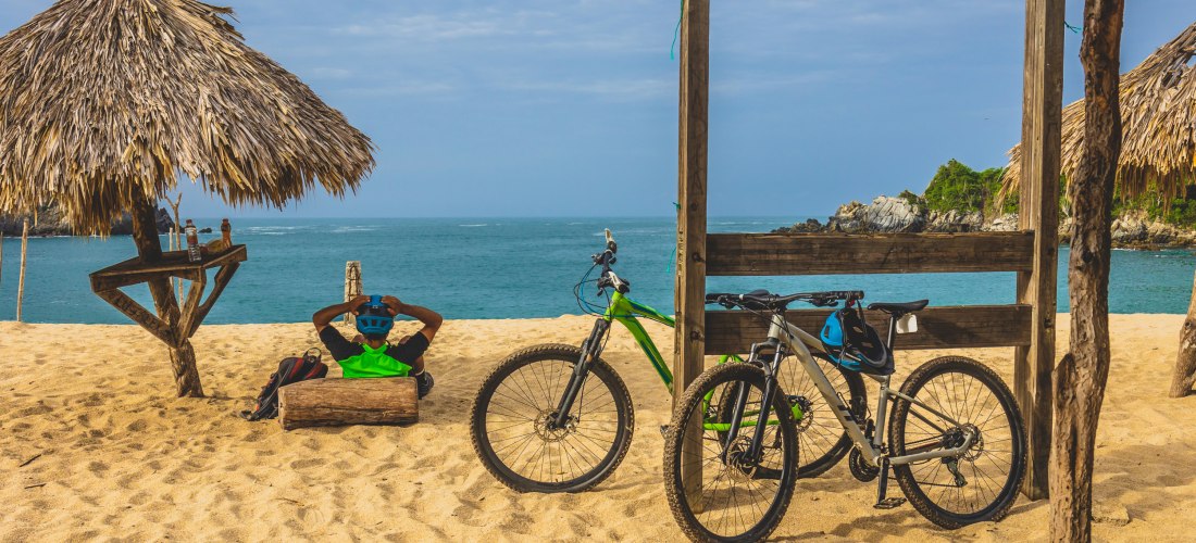 Zwei Mountainbikes im Sand neben einer Hütte mit Palmdach, während ein Radfahrer in neongrüner Weste entspannt am Strand sitzt und aufs Meer blickt