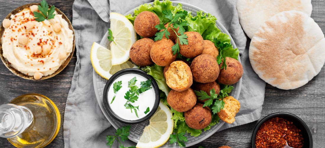 Ein Teller mit knusprigen Falafelbällchen auf Salat, serviert mit Zitronenscheiben, Joghurt-Dip, Fladenbrot und Hummus. Dazu Gewürze und eine Flasche Olivenöl auf einem dunklen Holztisch