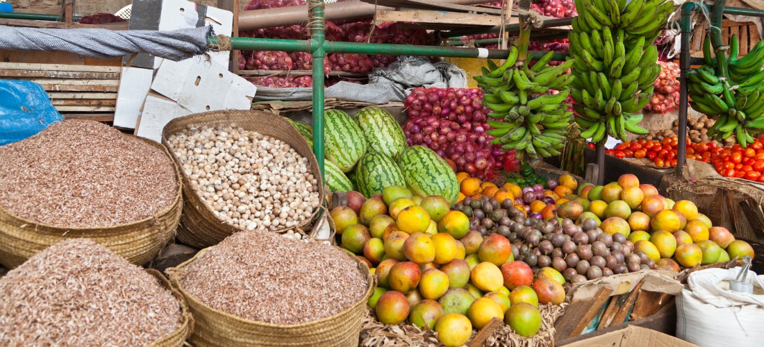 Ein farbenfroher Marktstand in Kenia mit einer Vielfalt an Lebensmitteln: Körbe mit getrockneten kleinen Fischen, Knoblauchknollen, Wassermelonen, Mangos, Passionsfrüchten, Bananen, Zwiebeln, Tomaten und anderen Produkten. Alles liebevoll aufgestapelt.