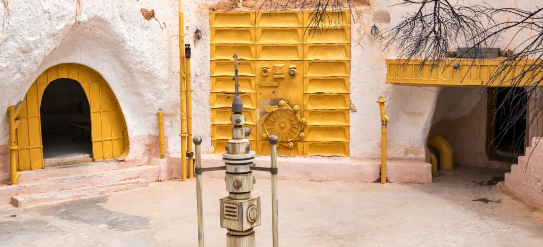Innenhof eines traditionellen Höhlenhauses in Matmata, Tunesien. Der Boden ist aus gestampftem Lehm, umgeben von gelb gestrichenen Türen, Torbögen und Felswänden. Gelbe Rohre und Lüftungselemente stechen aus der Wand hervor. Eine detaillierte Szenerie im 