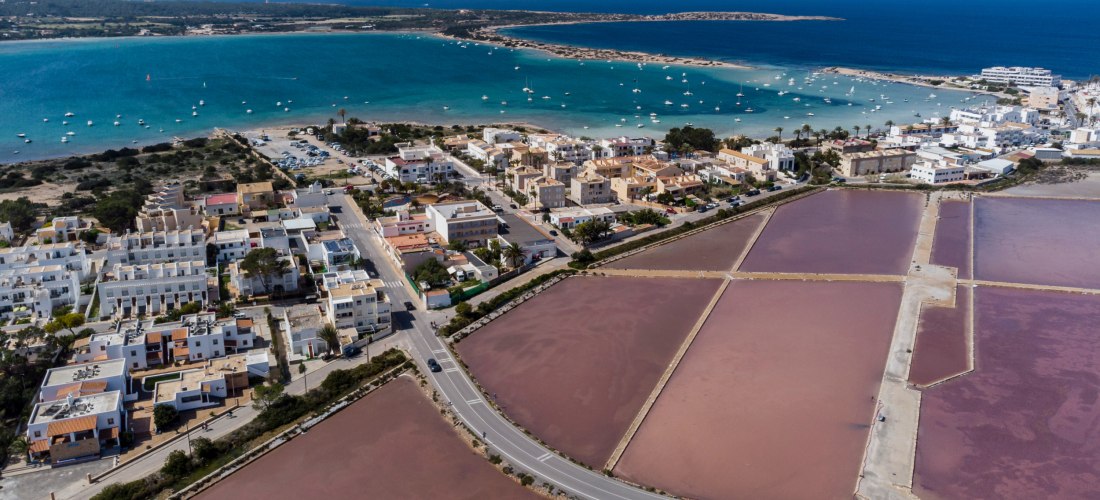Luftaufnahme von La Savina auf Formentera mit Hafen, türkisblauem Meer und vielen Booten. Im Vordergrund liegen die rosa schimmernden Salzbecken, dahinter die Küstenstadt mit weißen und beigefarbenen Häusern.