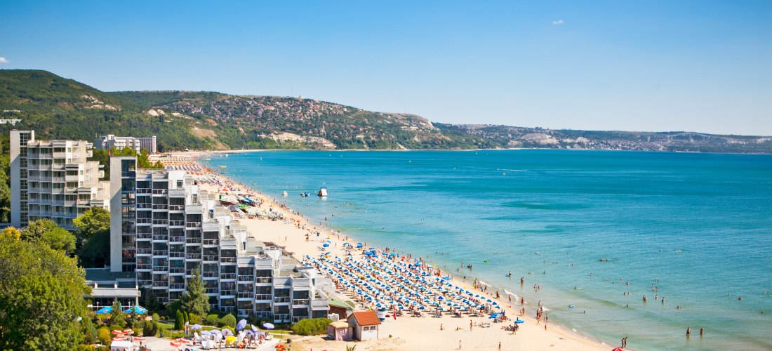 Blick auf ein belebtes Strandresort mit modernen Hotels an einem langen Sandstrand. Viele Sonnenschirme und Badende verteilen sich am türkisfarbenen Meer. Im Hintergrund grüne Hügel