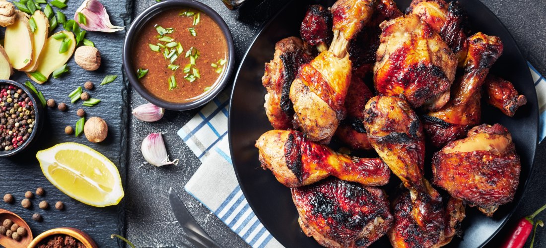 Ein schwarzer Teller voller gegrillter Jerk Chicken Drumsticks, die außen knusprig und kräftig gewürzt sind. Daneben liegen auf einer Schieferplatte frische Zutaten wie Ingwer, Knoblauch, Zitronenspalten, Chili, Pfefferkörner, Muskatnuss und brauner Zucke