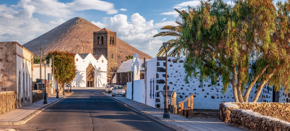 Ruhige Straße mit weiß getünchten Häusern und Palmen, im Hintergrund eine historische Kirche mit Glockenturm und ein kegelförmiger Vulkan unter blauem Himmel mit Wolken.