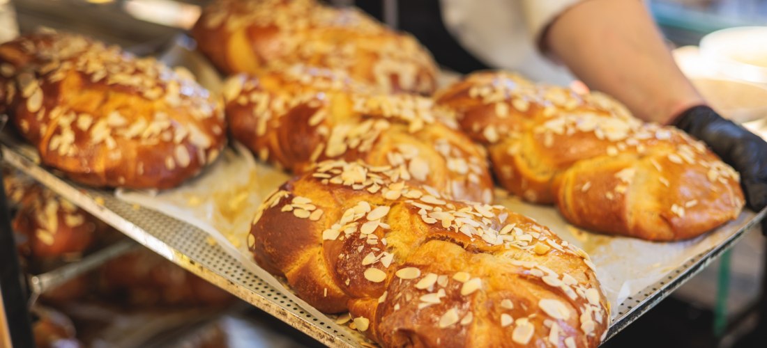 Frisch gebackene, goldbraune Hefezöpfe auf einem Backblech, bedeckt mit gehobelten Mandeln. Ein Bäcker mit weißem Hemd und Schürze hält das Blech in der Hand. Das Gebäck sieht fluffig und glänzend aus, bereit zum Verkauf oder Servieren