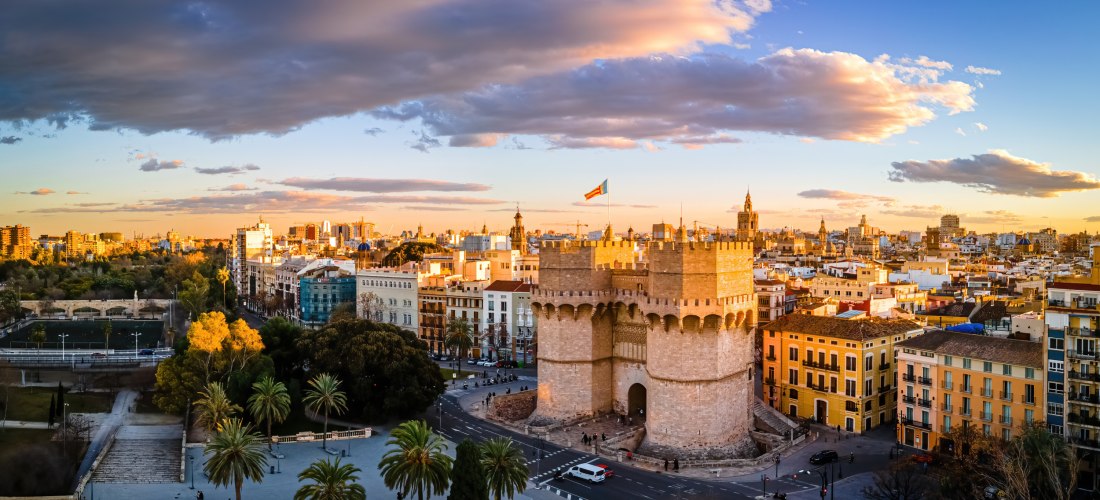 Panoramablick auf Valencia bei Sonnenuntergang. Im Vordergrund das imposante Stadttor Torres de Serranos mit wehender Flagge. Umgeben von Palmen, Altstadtgebäuden und dem ehemaligen Flussbett des Turia-Parks.