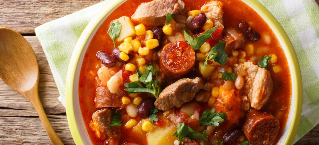Nahaufnahme eines kapverdischen Eintopfs in einem tiefen Teller. Der Eintopf enthält Fleischstücke, Chorizo-Wurst, Mais, Bohnen, Kartoffeln und Tomaten in einer roten Brühe, garniert mit frischer Petersilie. Daneben liegt ein Holzlöffel auf einem karierte