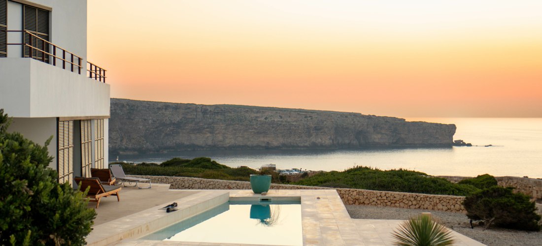 Moderne weiße Villa mit Poolterrasse auf Menorca bei Sonnenuntergang. Der Infinity-Pool spiegelt den Himmel in zarten Orange- und Gelbtönen wider. Im Hintergrund erhebt sich eine flache Steilküste über dem ruhigen Meer. Umgeben ist die Anlage von Steinmau