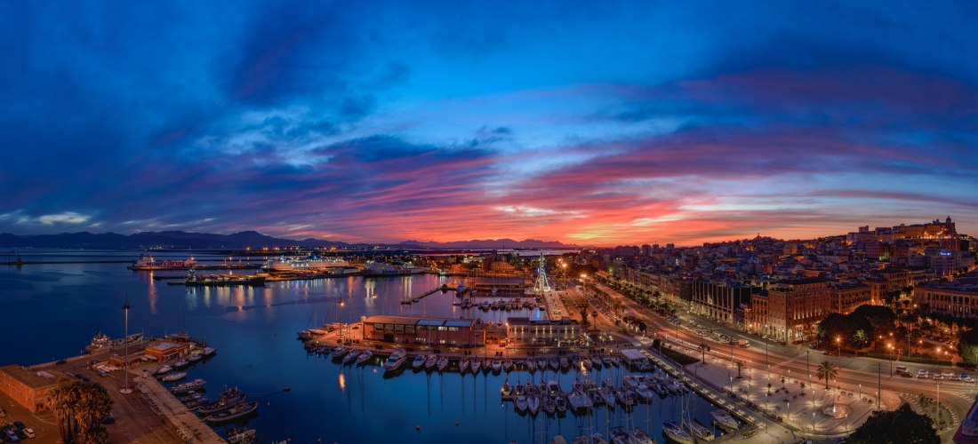 Panoramablick auf den Hafen von Cagliari bei Sonnenuntergang mit buntem Himmel, Lichtern der Stadt und Yachten im Mittelmeer – Hauptstadt Sardinien