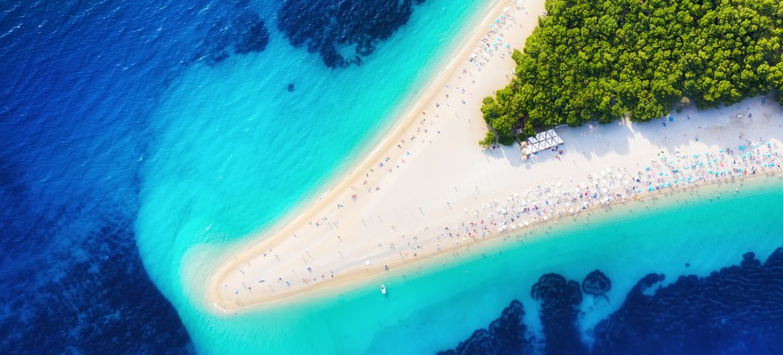 Draufsicht auf den berühmten Strand Zlatni Rat auf der kroatischen Insel Brač. Die schmale, sichelförmige Landzunge mit hellem Kiesstrand ragt ins türkisfarbene Meer, umgeben von dichtem Pinienwald und Badegästen mit bunten Sonnenschirmen.