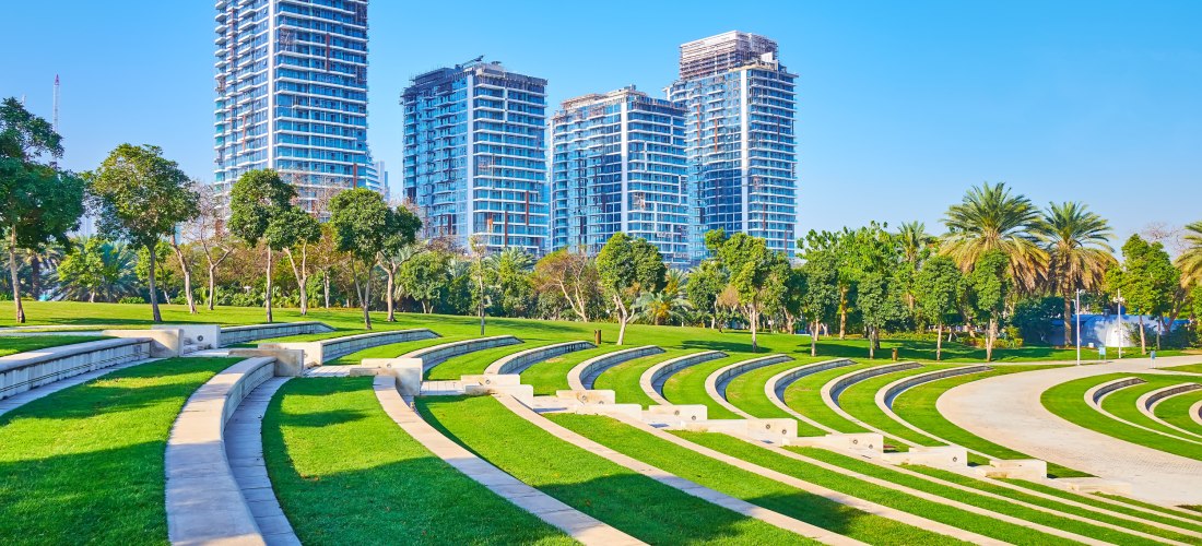 Dubai - Zabeel Park Modern gestaltete Grünanlage mit runden Terrassen aus Rasen und Stein, die an ein Amphitheater erinnern. Im Hintergrund ragen mehrere moderne Hochhäuser in den Himmel. Palmen und Bäume säumen den Park, der Himmel ist klar und blau.