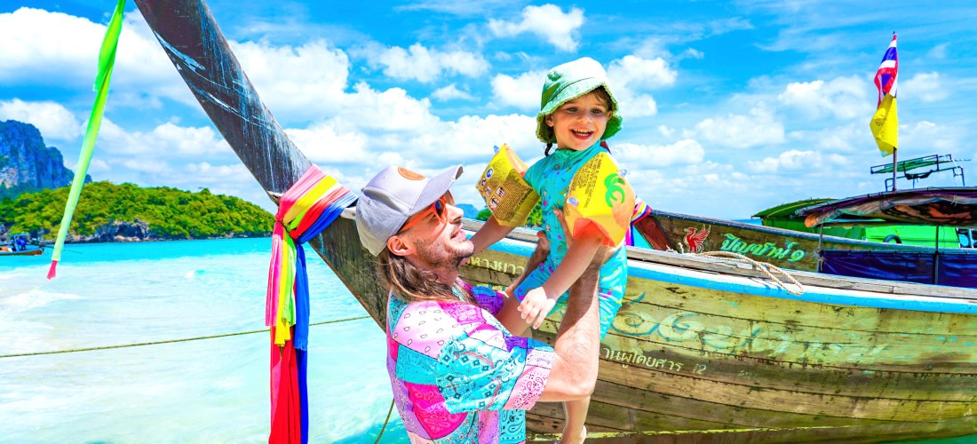 Das Bild zeigt einen Vater, der sein kleines Kind lachend in die Luft hebt. Beide stehen im seichten, türkisfarbenen Wasser vor einem traditionellen thailändischen Langboot, das mit bunten Stoffbändern und Flaggen geschmückt ist. Der Vater trägt ein farbe