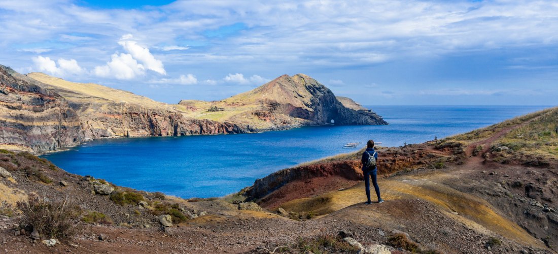 Person mit Rucksack blickt auf eine zerklüftete Küste mit türkisblauem Wasser auf Madeira. Der Wanderweg führt über farbige Felsen und Hänge, während sich im Hintergrund eine markante Landzunge ins Meer erstreckt. Der Himmel ist leicht bewölkt, aber freun