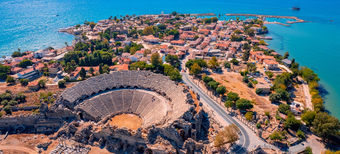 Das Bild zeigt eine Luftaufnahme von Side an der türkischen Riviera. Im Vordergrund liegt ein gut erhaltenes römisches Amphitheater, dahinter eine Altstadt mit roten Dächern auf einer schmalen Landzunge im Mittelmeer. Ruinen, enge Gassen und ein kleiner H