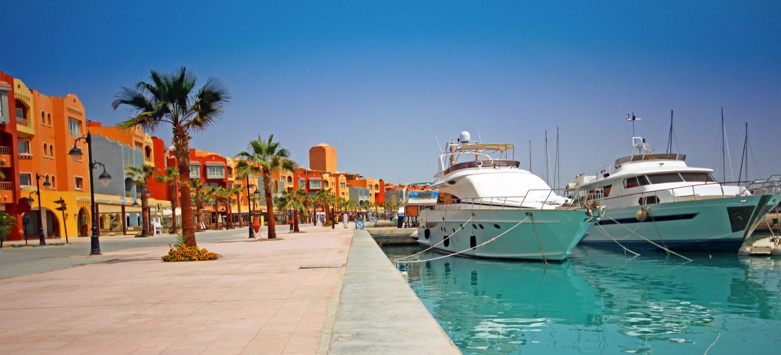 Das Bild zeigt die lebendige Marina von Hurghada am Roten Meer. Rechts im Bild liegen mehrere luxuriöse Yachten und Boote im türkisfarbenen Wasser vertäut. Links verläuft eine breite, gepflegte Promenade mit Palmen, Blumenbeeten und farbenfrohen Gebäuden 